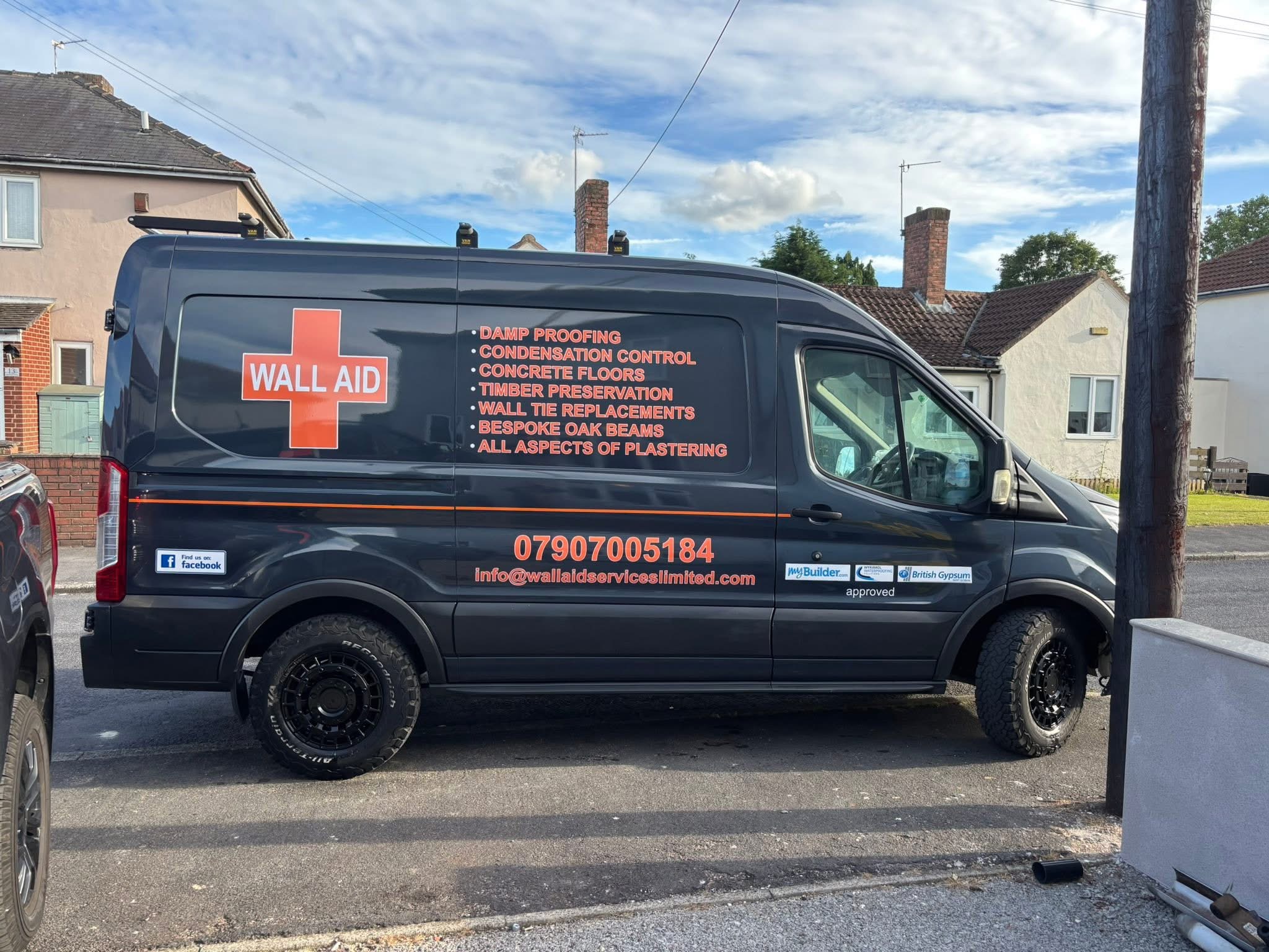 The Wall Aid van, a dark grey Transit with the orange cross logo, a painted list of services, the phone number and email