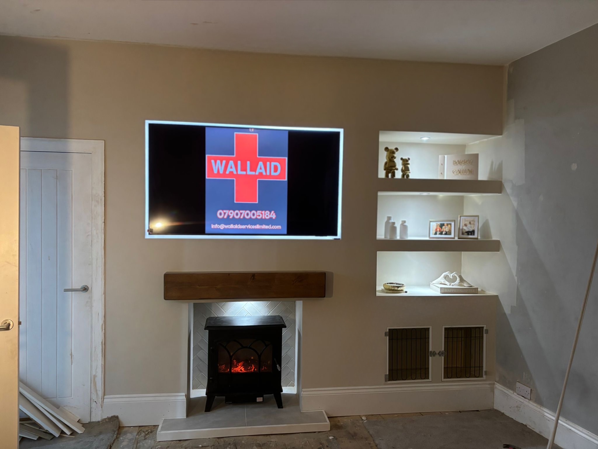 A finished media wall: TV set into the wall showing the Wall Aid logo, a timber beam over a stove fireplace and lit alcove shelves